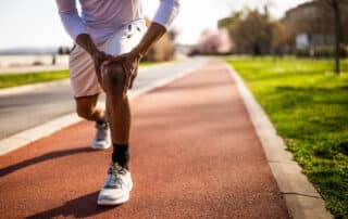 Male athlete clutching his knee in pain while jogging on an outdoor track, highlighting a potential sports injury that may require sports medicine treatment