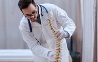 An orthopedic surgeon for spinal fusion in white uniform and with stethoscope around neck holding spine model while standing in hospital.