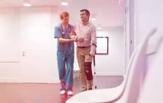 Nurse assisting patient with injured leg in walking at walk-in orthopedic urgent care corridor.